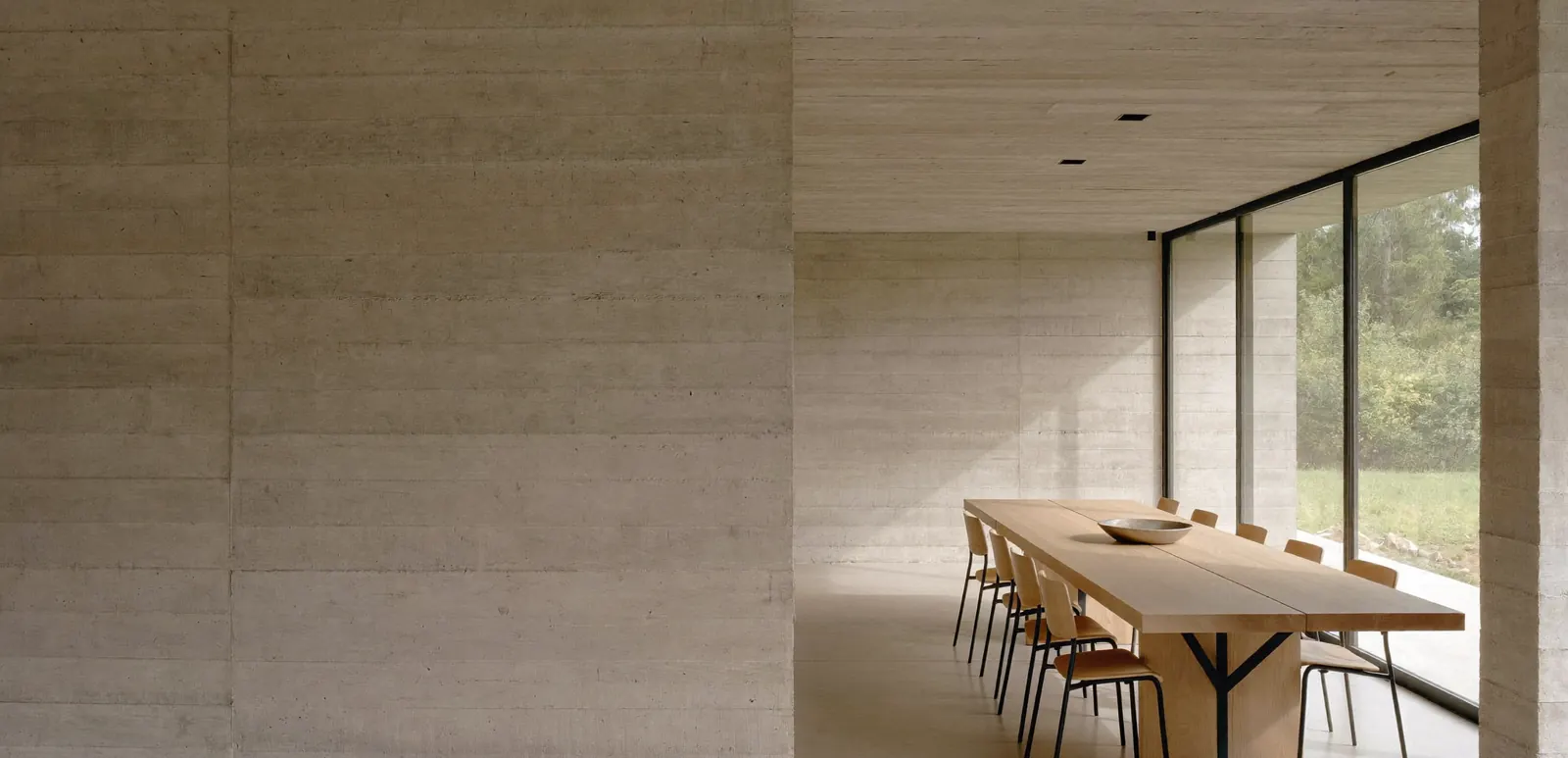 Comedor en una casa de hormigón con mesa larga de roble, sillas de madera y ventanal con estructura de acero